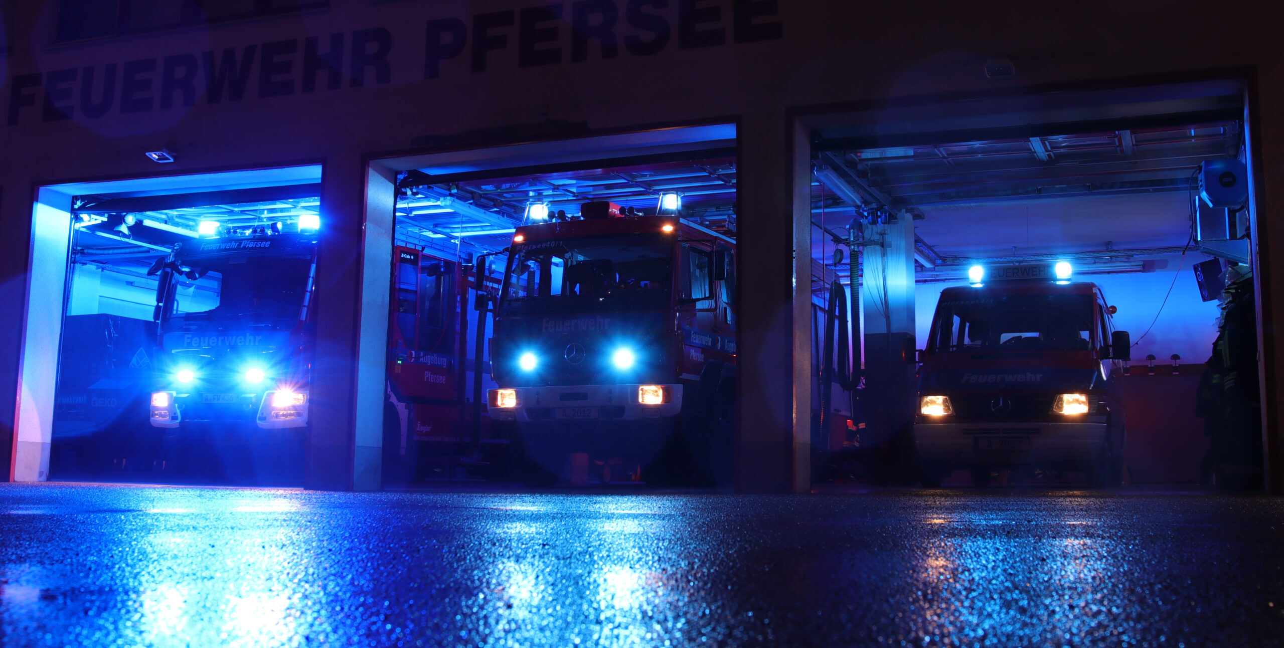 Fahrzeughalle der Feuerwehr Pfersee bei Nacht - alle drei Fahrzeuge mit Blaulicht zu sehen. 